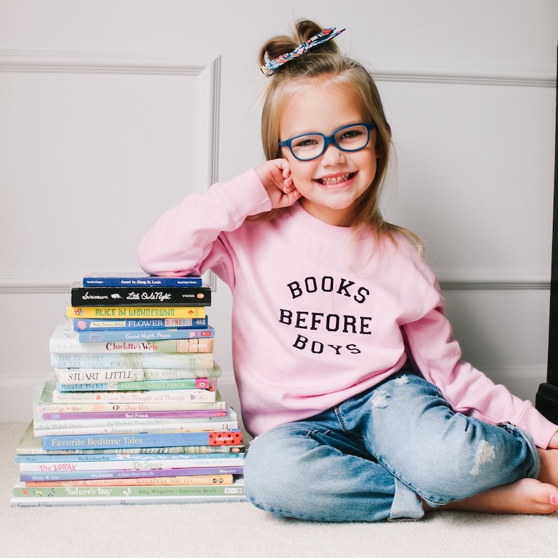 May include: A young girl wearing a pink sweatshirt with the text "BOOKS BEFORE BOYS" in black lettering. She is sitting on a carpeted floor with a stack of children's books in front of her.