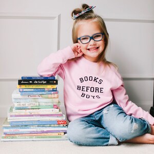 May include: A young girl wearing blue jeans and a pink sweatshirt with the text "BOOKS BEFORE BOYS" is sitting on a light brown surface. She is holding a stack of books in front of her. The books include titles such as "Alice in Wonderland", "Charlotte's Web", and "Stuart Little".