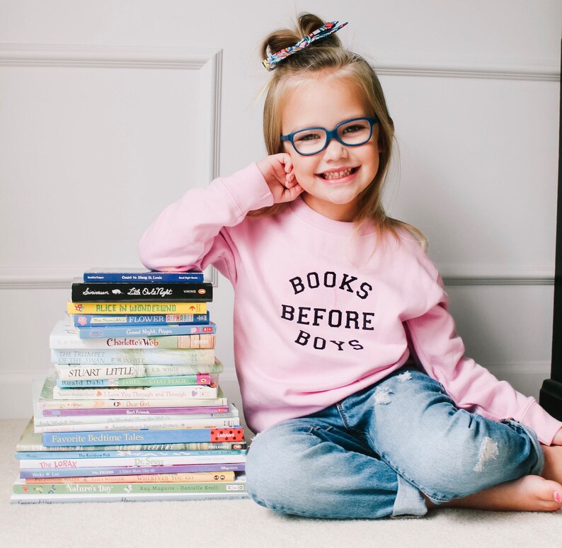 May include: A young girl wearing blue jeans and a pink sweatshirt with the text "BOOKS BEFORE BOYS" is sitting on a light brown surface. She is holding a stack of books in front of her. The books include titles such as "Alice in Wonderland", "Charlotte's Web", and "Stuart Little".