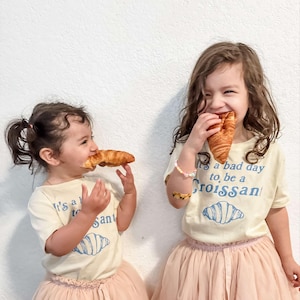 May include: Two young girls wearing white t-shirts with a blue graphic of a croissant and the text "It's a bad day to be a Croissant". One girl is wearing a pink tulle skirt and the other is wearing a pink tulle skirt with a white lace trim. Both girls are holding and eating croissants.