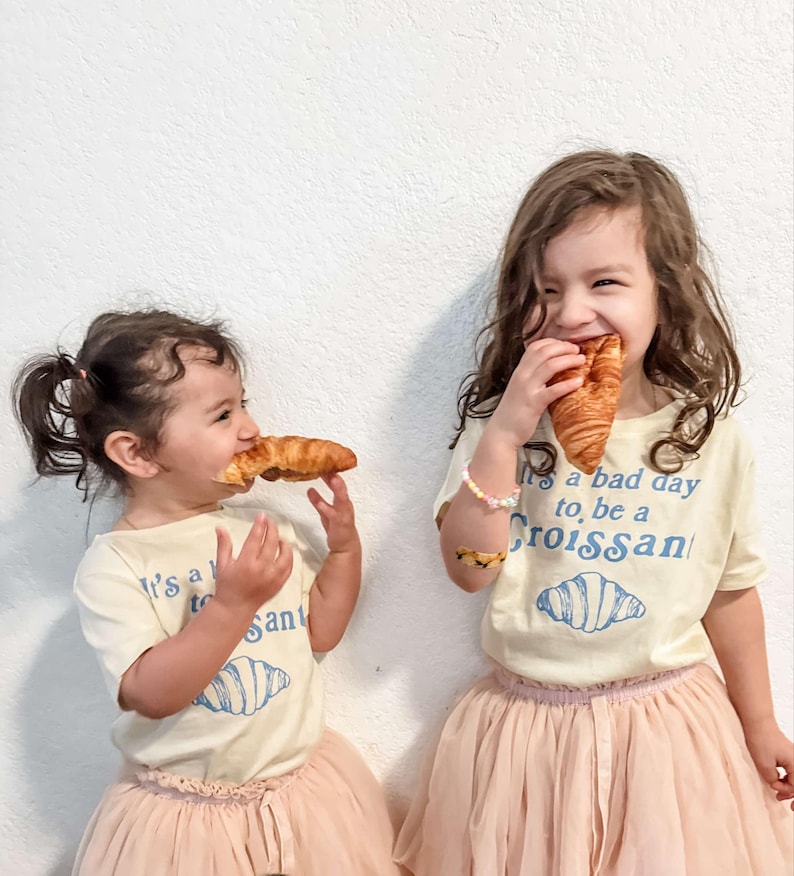 May include: Two young girls wearing white t-shirts with a blue graphic of a croissant and the text "It's a bad day to be a Croissant". One girl is wearing a pink tulle skirt and the other is wearing a pink tulle skirt with a white lace trim. Both girls are holding and eating croissants.