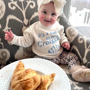 May include: A baby wearing a white headband and a light beige sweatshirt with the text "It's a bad day to, be a Croissant" in blue. The baby is sitting on a gray and white patterned chair with a croissant on a white plate in front of them.