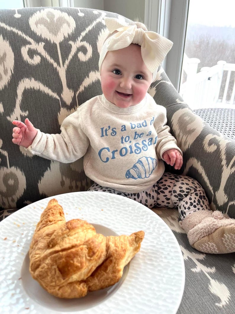 May include: A baby wearing a white headband and a light beige sweatshirt with the text "It's a bad day to, be a Croissant" in blue. The baby is sitting on a gray and white patterned chair with a croissant on a white plate in front of them.