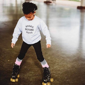 May include: A young person wearing a grey sweatshirt with the text "BOOKS BEFORE BOYS" in black lettering. They are wearing black pants and pink and white striped socks. They are roller skating on black and gold roller skates.
