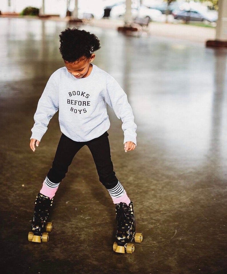 May include: A young person wearing a grey sweatshirt with the text "BOOKS BEFORE BOYS" in black lettering. They are wearing black pants and pink and white striped socks. They are roller skating on black and gold roller skates.