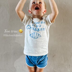 May include: A young child wearing a white t-shirt with blue text that reads "It's a bad day to be a Croissant" and a blue illustration of a croissant. The child is holding a croissant above their head and is wearing blue shorts and blue and white striped socks.