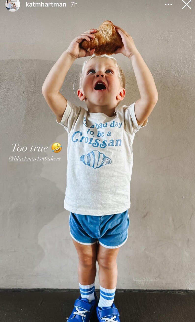 May include: A young child wearing a white t-shirt with blue text that reads "It's a bad day to be a Croissant" and a blue illustration of a croissant. The child is holding a croissant above their head and is wearing blue shorts and blue and white striped socks.