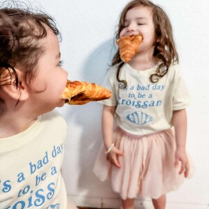 May include: Two young children wearing white t-shirts with the text "It's a bad day to be a croissant" in blue lettering. The children are both eating croissants. The child on the left is wearing a pink tulle skirt. The child on the right is wearing a pink tulle skirt.
