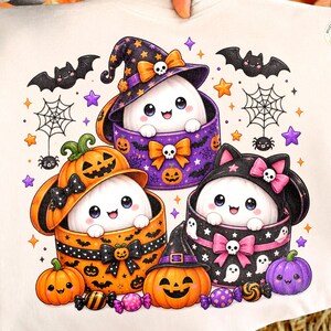 Peut inclure: Un t-shirt couleur crème avec une illustration de trois personnages blancs fantomatiques sortant de boîtes décorées. L'un est dans une boîte violette sur le thème des citrouilles, un autre dans une citrouille orange et le dernier dans une boîte noire.