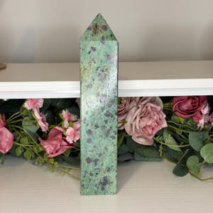 May include: A rectangular crystal obelisk with a pointed top. The obelisk is a mottled green colour with dark purple and red inclusions. The crystal is displayed on a white shelf with pink and green floral accents.