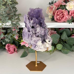 May include: A large amethyst geode cluster in shades of lavender and white, mounted on a gold-coloured metal stand. The geode has a textured, bubbly surface. It is displayed against a backdrop of flowers and greenery.