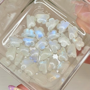 May include: A selection of small, cloud-shaped moonstone carvings in a clear, square container. The carvings are white with iridescent blue and yellow flashes. The container is held by a person.