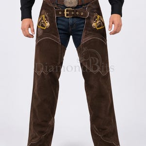 May include: Dark brown suede leather chaps featuring yellow embroidered horse head designs on the upper thighs, a front belt closure with a brass buckle, and decorative stitching along the leg seams. The text Diamond Bits is visible.