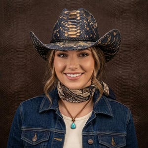 Può includere: Un cappello da cowboy con motivo a pelle di serpente blu scuro e marrone chiaro, dotato di una fascia intrecciata nera. Il cappello presenta una corona strutturata e una tesa curva. La superficie mostra una texture simile a delle squame.