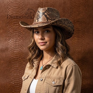 Puede incluir: Un sombrero de vaquero con un patrón de piel de serpiente marrón y bronceado con una banda trenzada alrededor de la corona. El sombrero se coloca sobre una chaqueta de mezclilla bronceada y un collar con colgante turquesa. El texto DiamondBits es visible.