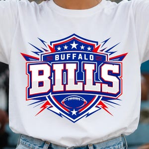Puede incluir: Una camiseta blanca de manga corta con el logotipo de los Buffalo Bills. El diseño incluye un escudo azul con detalles en rojo, estrellas blancas, el texto BUFFALO BILLS en letras blancas en negrita y una ilustración de un balón de fútbol.