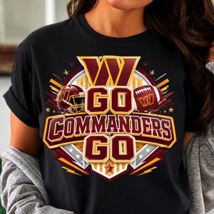May include: A black t-shirt featuring a graphic design with the text W GO COMMANDERS GO in burgundy and gold. The design includes a football helmet and a football, surrounded by star and lightning bolt accents on a circular background.