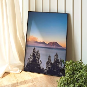 Könnte beinhalten: Gerahmter Landschaftsdruck mit einer Bergkette bei Sonnenuntergang. Das Bild zeigt einen lebendigen Himmel mit Blautönen, Orange und Rosa. Silhouetten von Bäumen befinden sich im Vordergrund. Der Druck steht auf einem Holzboden.