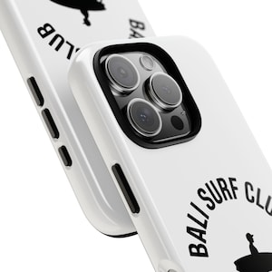 May include: White phone case with black accents and a black and white graphic. The case features the text "BALI SURF CLUB" in a circular design, along with a silhouette of a surfer. The phone case is designed to protect the phone.