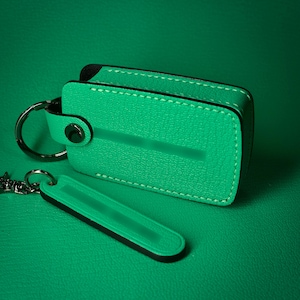 May include: A green leather key holder and key fob set. The key holder is rectangular with black trim and white stitching. The key fob is a smaller, elongated rectangle. Both items are on a green surface.