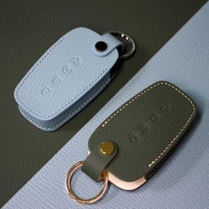 Op de afbeelding: Twee lederen sleuteletuis met een generfde textuur, één lichtblauw en één olijfgroen. Elk etui is voorzien van een drukknoopsluiting, een metalen sleutelring en een reliëfopdruk op de voorzijde. De etuis hebben een rechthoekige vorm.