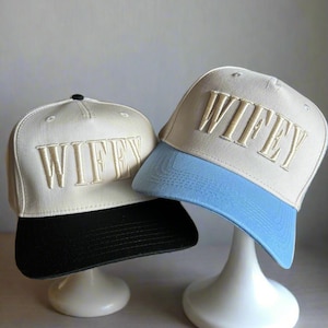 May include: Two beige baseball caps with black and blue brims. The caps have the word "WIFEY" embroidered in gold on the front.