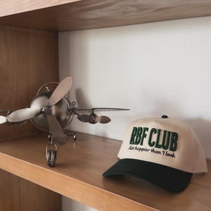Könnte beinhalten: Eine beige Baseballkappe mit grünem Schirm und dem gestickten Text "RBF CLUB  A lot happier than I look" in Grün.