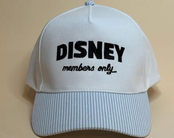 May include: White baseball cap with the word "DISNEY" in bold black letters, and "members only" in a script font. The brim features light blue and white vertical stripes. A small, striped button is on top.