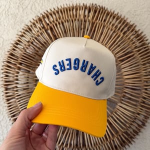Könnte beinhalten: Eine cremefarbene und gelbe Baseballkappe mit dem Wort "CHARGERS" in Blau gestickt. Die Kappe hat einen gelben Schirm und einen gelben Knopf oben. Die Kappe wird von einer Hand gehalten.