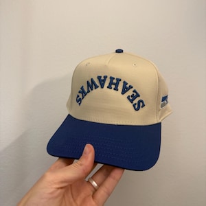 May include: A beige and blue baseball cap with the word "SEAHAWKS" embroidered in blue across the front. The cap has a blue brim and a small blue button on top. A small white and silver tag is on the side.