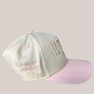 May include: A white and pink baseball cap with the text "let me do your hair" embroidered in pink on the front. The cap has a white crown and a pink brim.