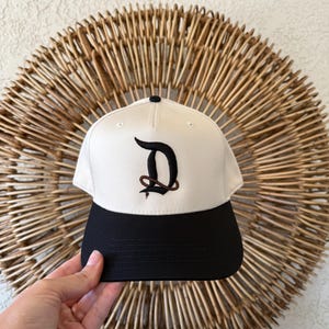 May include: A baseball cap with a cream-colored crown and a black brim. The cap features a stylized, embroidered letter "D" in dark brown. The cap has a black button on top.