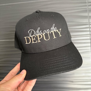 Può includere: Cappellino da baseball nero con la scritta ricamata "Dibs on the DEPUTY" in bianco e oro. Il cappellino ha una visiera curva e un bottone sulla parte superiore. Un accessorio casual.
