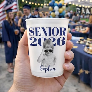 May include: A white plastic cup with the text "SENIOR 2026" in navy blue. A black and white photo of a young girl wearing sunglasses is below the text, with the name "Sophia" beneath the photo. The cup is held in a hand at a graduation party.