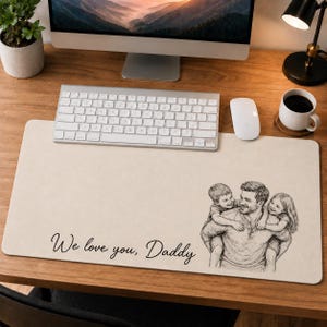 May include: A beige desk mat featuring a black and white illustration of a father with two children. The cursive text reads "We love you, Daddy". A keyboard, mouse, and a coffee cup are visible on the wooden desk.