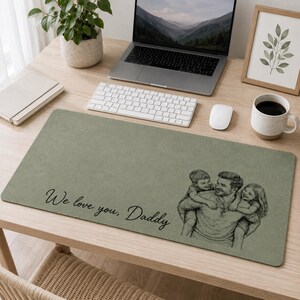 May include: A rectangular sage green desk mat featuring the text "We love you, Daddy" and a sketch of a father with two children. The mat is on a light wood desk, with a laptop, keyboard, and a coffee mug.