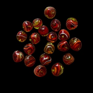 May include: A collection of twenty small, round glass beads featuring a swirling pattern of deep red, orange, and white colors. Each bead has a smooth, glossy finish with translucent and opaque sections, creating a marbled effect across the set.