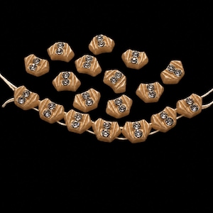 May include: A collection of small, gold-toned, fan-shaped buttons featuring a textured, ribbed surface and two embedded clear, circular rhinestones in the center. Some buttons are loose, while others are strung together on a thin, light-colored cord.
