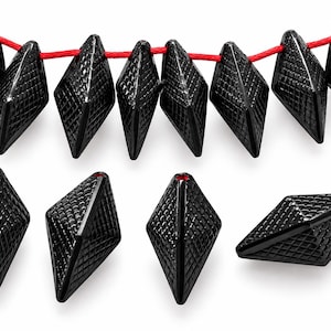 May include: A collection of black diamond-shaped beads featuring a textured, cross-hatched grid pattern on their surfaces. Several beads are strung together on a bright red cord, while four individual beads are displayed separately below.