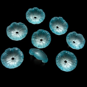 Puede incluir: Un conjunto de siete cuentas de color azul claro, translúcidas y con forma de flor. Cada cuenta presenta un borde festoneado, un orificio central y un patrón de pétalos radiantes texturizados. Una cuenta está de lado, las otras planas.