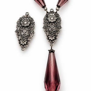 May include: A jewelry component set featuring two ornate, silver-toned metal filigree connectors with floral motifs and three faceted, deep purple teardrop-shaped crystals. The pieces are arranged with one connector separate and one attached to crystals.