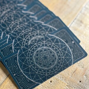 K&ouml;nnte beinhalten: Ein Stapel Spielkarten mit dunkelblauem Hintergrund und silbernem Design. Das Design zeigt einen Kreis mit einem Stern in der Mitte und geometrischen Mustern.