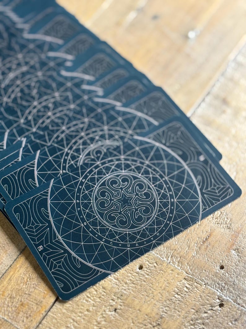 K&ouml;nnte beinhalten: Ein Stapel Spielkarten mit dunkelblauem Hintergrund und silbernem Design. Das Design zeigt einen Kreis mit einem Stern in der Mitte und geometrischen Mustern.