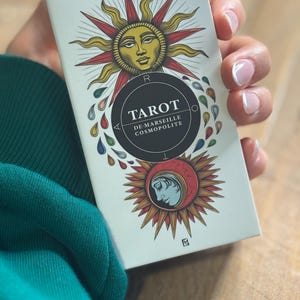 K&ouml;nnte beinhalten: Eine wei&szlig;e Schachtel mit einem schwarzen Kreis, der den Text "Tarot de Marseille Cosmopolite" enth&auml;lt. Die Schachtel zeigt eine farbenfrohe Illustration einer Sonne mit einem Gesicht und eines zunehmenden Mondes mit einem Gesicht.