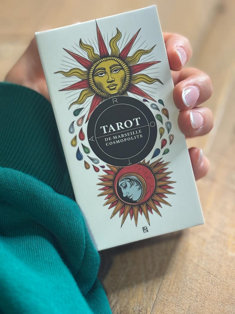 K&ouml;nnte beinhalten: Eine wei&szlig;e Schachtel mit einem schwarzen Kreis, der den Text "Tarot de Marseille Cosmopolite" enth&auml;lt. Die Schachtel zeigt eine farbenfrohe Illustration einer Sonne mit einem Gesicht und eines zunehmenden Mondes mit einem Gesicht.