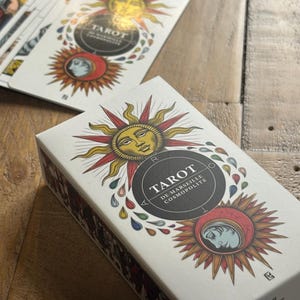 K&ouml;nnte beinhalten: Eine wei&szlig;e Schachtel mit einem goldenen und roten Sonnenmotiv. Auf der Vorderseite der Schachtel steht "Tarot De Marseille Cosmopolite". Die Schachtel liegt auf einer Holzoberfl&auml;che.