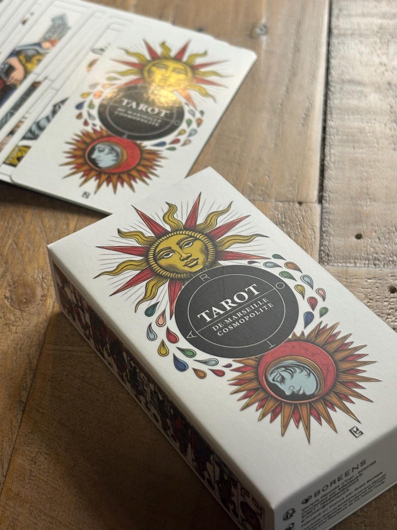 K&ouml;nnte beinhalten: Eine wei&szlig;e Schachtel mit einem goldenen und roten Sonnenmotiv. Auf der Vorderseite der Schachtel steht "Tarot De Marseille Cosmopolite". Die Schachtel liegt auf einer Holzoberfl&auml;che.