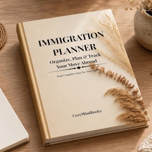 May include: A hardcover book titled "IMMIGRATION PLANNER" with text that reads "Organize, Plan & Track Your Move Abroad." The book is beige with gold trim and sits on a wooden surface. A cup of coffee, a notepad, and glasses are also visible.
