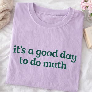 May include: A lilac-colored t-shirt with the text "it's a good day to do math" printed in dark green. The shirt is folded neatly on a white surface. The image also includes a glimpse of a beige sweater, flowers, and a book.