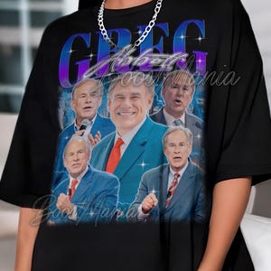 Puede incluir: Camiseta negra con un gráfico de un hombre con traje, el nombre "GREG" en letras grandes y estilizadas y el texto "Abbott Boost Mania" en cursiva.